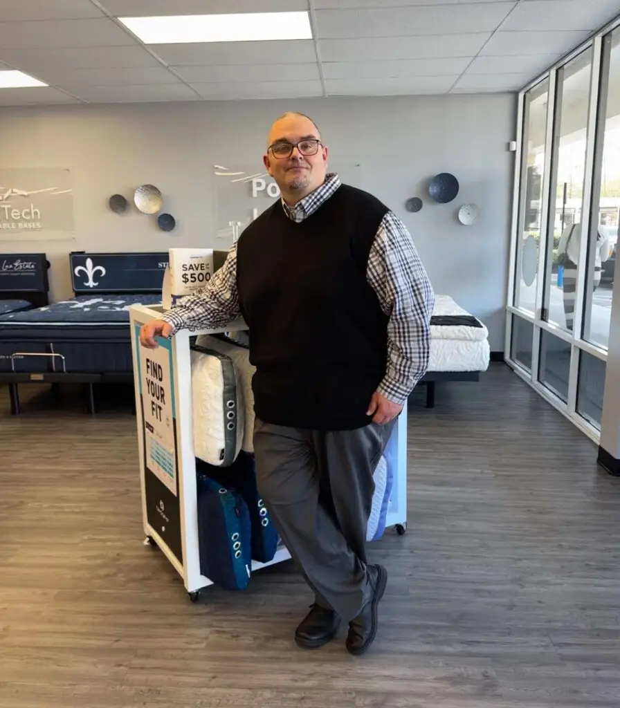 Man standing beside a display of mattresses and bedding accessories in a Jacksonville Bedding store, promoting the "Find Your Fit" mattress matching service.