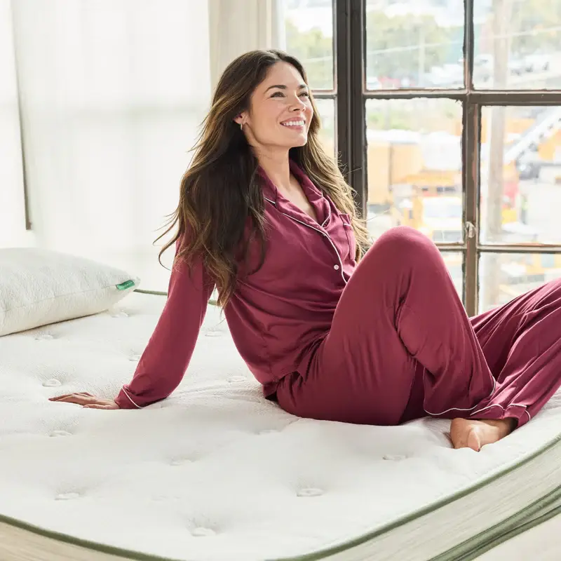 Woman in maroon pajamas smiling while sitting on Avocado Green Mattress Box Top, showcasing comfort and luxury in a bright, airy bedroom setting.