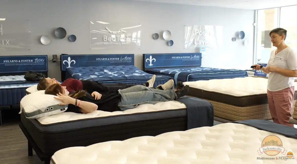Two individuals lying on a mattress in a showroom, exploring comfort options, with a sales associate standing nearby, discussing mattress features from Stearns & Foster.