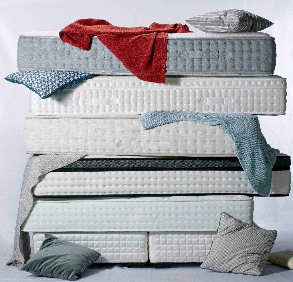 Stacked mattresses in various thicknesses and textures, featuring decorative pillows and blankets, illustrating customization and comfort for diverse sleeper types.