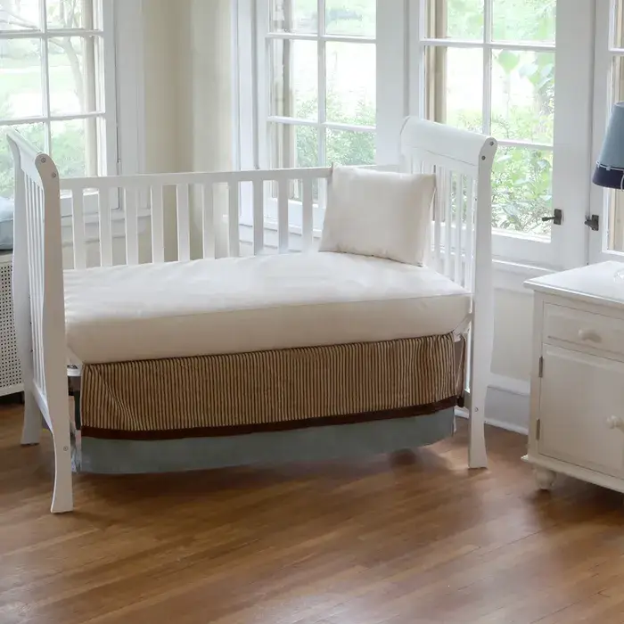 Crib mattress in a white crib with a striped skirt, positioned in a bright room with large windows, symbolizing safe sleep for babies, part of Jacksonville Bedding's initiative to support families affected by wildfires.